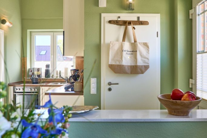 Grüne Küche mit Wandregal, Tasche an der Tür und Obstschale auf der Theke.